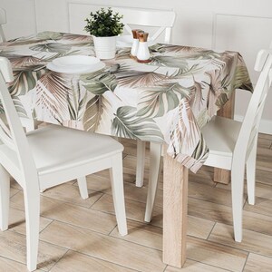 Könnte beinhalten: Ein Esstisch-Set mit einer weißen Tischdecke mit einem tropischen Blättermuster in Grün-, Braun- und Beigetönen. Der Tisch ist mit weißen Tellern, Salz- und Pfefferstreuern und einer kleinen Topfpflanze gedeckt. Weiße Stühle umgeben den Tisch.