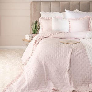 Peut inclure: Un lit avec une couette matelassée rose clair et des taies d'oreiller assorties à volants. Le lit est composé d'oreillers blancs et d'une couverture blanche. Une petite table de chevet contient une plante et un objet décoratif.