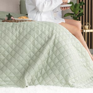 Könnte beinhalten: Eine hellgrüne, gesteppte Tagesdecke mit Rautenmuster. Die Tagesdecke liegt auf einem Bett mit einem weißen, flauschigen Teppich am Fußende. Eine Person sitzt auf dem Bett und trägt ein weißes Hemd. Ein Tablett mit Essen und Blumen befindet sich auf dem Bett.