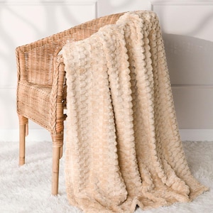 Könnte beinhalten: Eine beige strukturierte Decke, die über einem Korbstuhl drapiert ist. Die Decke hat ein weiches, plüschiges Aussehen mit einem sich wiederholenden Muster. Der Stuhl besteht aus geflochtenem Korb und hat eine natürliche, hellbraune Farbe. Die Decke liegt auf einem weißen, flauschigen Teppich.