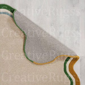 May include: Beige rug with a green, blue, and gold border. The rug's underside is visible, revealing a grey backing. The rug has a wavy design and is partially rolled up, showcasing its texture and colour variations.