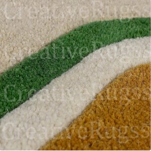 May include: Close-up of a rug with a rainbow design. The rug features curved stripes in beige, green, white, and gold. The texture of the rug appears soft and plush, with a dense pile. The design is simple and modern.