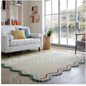 May include: A cream-coloured, rectangular rug with a wavy, scalloped border. The border features bands of green, blue, yellow, and rust colours. The rug is in a living room with a white sofa and large windows.