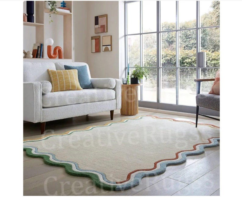 May include: A cream-coloured, rectangular rug with a wavy, scalloped border. The border features bands of green, blue, yellow, and rust colours. The rug is in a living room with a white sofa and large windows.