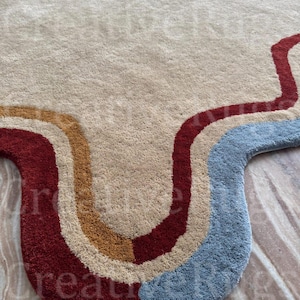 May include: A beige rug with a wavy design in red, tan, and blue. The rug has a soft, textured surface and a unique, retro-inspired pattern. The rug is suitable for a living room or bedroom.