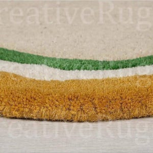 May include: Close-up of a round rug with a textured surface. The rug features a design with three curved bands in green, white, and gold. The rug is made of a soft, plush material.