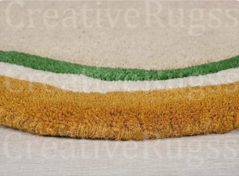 May include: Close-up of a round rug with a textured surface. The rug features a design with three curved bands in green, white, and gold. The rug is made of a soft, plush material.