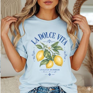 Może przedstawiać: Jasnoniebieski t-shirt z kr&oacute;tkim rękawem z grafiką przedstawiającą trzy ż&oacute;łte cytryny na gałązce z zielonymi liśćmi i białym kwiatem. Napis głosi LA DOLCE VITA, ITALIAN SUMMER, limoni di amalfi oraz EST. 1984 niebieskimi, ozdobnymi literami.