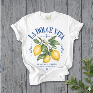 Może przedstawiać: Biały t-shirt z kr&oacute;tkim rękawem z grafiką przedstawiającą trzy ż&oacute;łte cytryny na ulistnionej gałązce z białymi kwiatami. Niebieski napis głosi LA DOLCE VITA, ITALIAN SUMMER, limoni di amalfi oraz EST. 1984, z ozdobnymi niebieskimi zawijasami.
