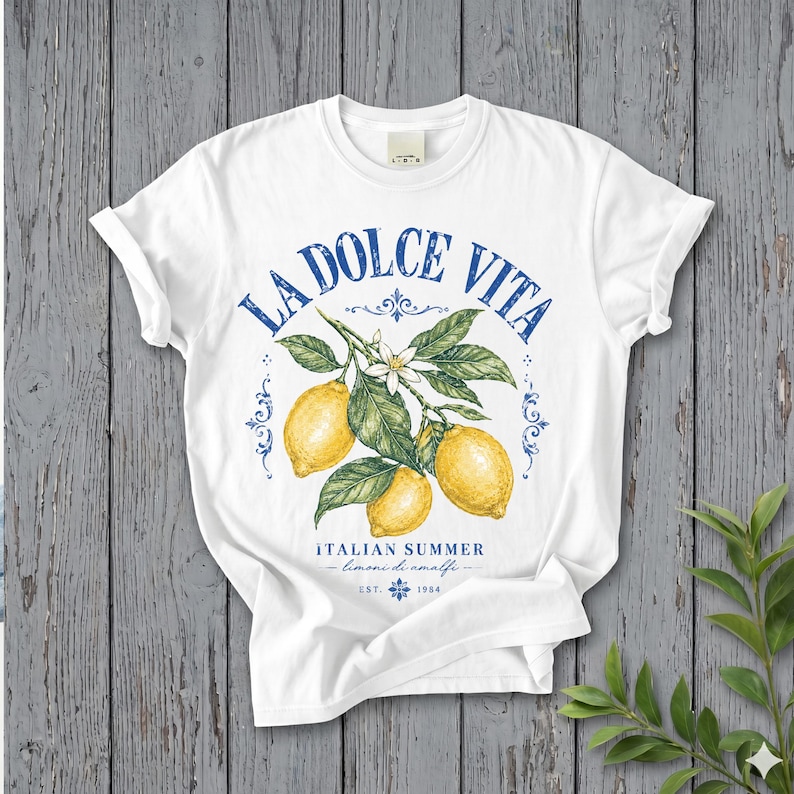 Może przedstawiać: Biały t-shirt z kr&oacute;tkim rękawem z grafiką przedstawiającą trzy ż&oacute;łte cytryny na ulistnionej gałązce z białymi kwiatami. Niebieski napis głosi LA DOLCE VITA, ITALIAN SUMMER, limoni di amalfi oraz EST. 1984, z ozdobnymi niebieskimi zawijasami.
