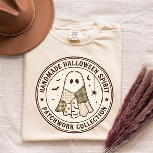 May include: A cream-coloured t-shirt featuring a circular graphic with the text "HANDMADE HALLOWEEN SPIRIT PATCHWORK COLLECTION." The design includes a ghost made of various green, beige, and white patterned patches, surrounded by small bats, a spider, and a crescent moon.