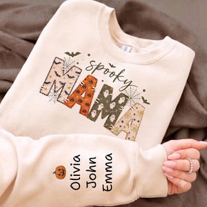 Può includere: Una felpa girocollo color crema con la scritta "spooky MAMA" in un carattere stile patchwork con motivi di Halloween come pipistrelli, zucche, fantasmi e ragnatele. La manica è personalizzata con un'icona di zucca e i nomi Olivia, John, Emma.