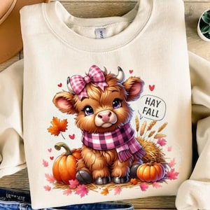 Puede incluir: Sudadera crema con una vaca de las Tierras Altas de dibujos animados con un lazo y bufanda de cuadros rosas, rodeada de calabazas, hojas y corazones. El texto "HAY FALL" aparece en una burbuja. Un diseño otoñal encantador.