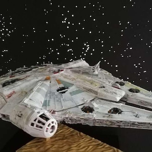 May include: A detailed model of the Millennium Falcon starship, featuring a white and gray color scheme with intricate paneling and detailing. The model is set against a black backdrop with white star-like dots, evoking a space theme.