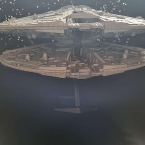May include: A detailed model of a spaceship, featuring a layered design with intricate details. The ship is primarily light gray, with a dark background and a reflective surface below, creating a mirrored effect. The model is mounted on a stand.