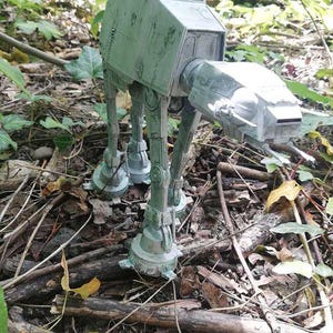Puede incluir: Un modelo detallado de un caminante AT-AT de Star Wars, en un color gris verdoso desgastado. El modelo está ambientado en un entorno natural de hojas y ramitas, mostrando su diseño mecánico.