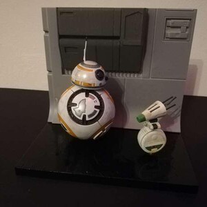 May include: A Star Wars BB-8 droid figure and a small, white and green droid figure are displayed on a black surface. The BB-8 figure is white with orange and black accents. A gray wall-like structure is in the background.