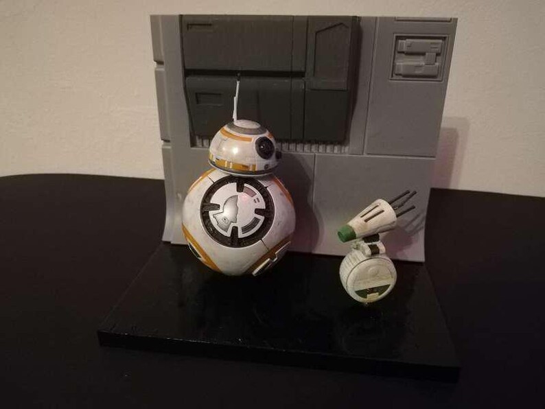 May include: A Star Wars BB-8 droid figure and a small, white and green droid figure are displayed on a black surface. The BB-8 figure is white with orange and black accents. A gray wall-like structure is in the background.