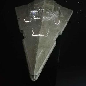 Puede incluir: Maqueta de una nave espacial Star Destroyer, con ventanas y detalles iluminados, sobre un fondo negro con estrellas blancas dispersas. La nave es de color gris claro y está montada sobre un soporte negro.