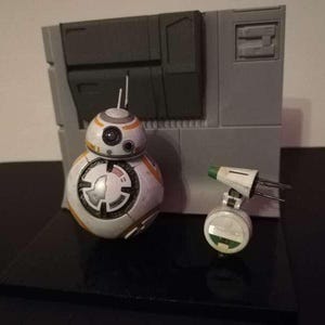 May include: A detailed model of BB-8 and a small white and green droid, set against a gray backdrop. BB-8 is white, orange, and silver, with a circular design. The small droid has a green top. The scene is set on a black base.