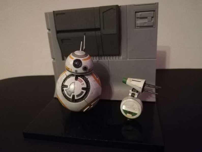 May include: A detailed model of BB-8 and a small white and green droid, set against a gray backdrop. BB-8 is white, orange, and silver, with a circular design. The small droid has a green top. The scene is set on a black base.
