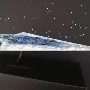 Könnte beinhalten: Modell eines Star Destroyer Raumschiffs, mit weißem und blauem Farbschema. Das Modell steht auf einem schwarzen Ständer vor einem schwarzen Hintergrund mit weißen sternartigen Punkten.