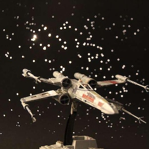 Op de afbeelding: Een model X-wing starfighter met rode markeringen en een wit en grijs kleurenschema, tegen een achtergrond van een sterrennacht. Het model is op een standaard gemonteerd.