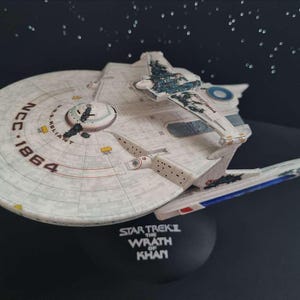 Puede incluir: Una maqueta de la nave estelar Star Trek Reliant, con una combinación de colores blanco y gris con detalles azules. La nave tiene un casco principal circular con el texto "NCC-1864" y "Reliant". La base dice "Star Trek II The Wrath of Khan."