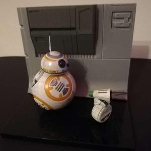 May include: A Star Wars BB-8 droid figure with a small companion droid, set against a gray backdrop. The BB-8 figure is white and orange, with black details. The small droid is white with green and black accents. The backdrop is gray.