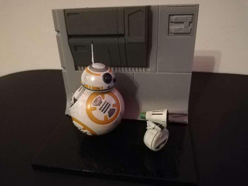 May include: A Star Wars BB-8 droid figure with a small companion droid, set against a gray backdrop. The BB-8 figure is white and orange, with black details. The small droid is white with green and black accents. The backdrop is gray.