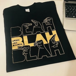 May include: Black t-shirt with a gold design featuring the words "BEAH BLAH BLAH" and hand gestures. The text "ARMIN VAN BUUREN" is also printed on the shirt. The shirt is made of a soft material.