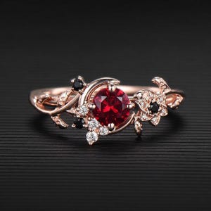May include: A rose gold ring featuring a central round-cut red gemstone, accented by smaller black stones and clear white stones. The band is designed with a vine-like motif, incorporating delicate leaf and floral shapes that wrap around the finger.