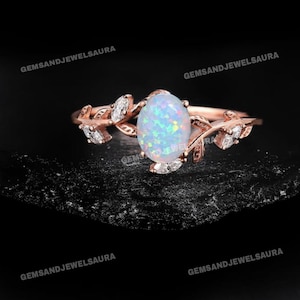 May include: Rose gold ring featuring an oval opal center stone with iridescent colors. The ring has a leaf design with small, clear accent stones. The ring is set against a dark, textured background.