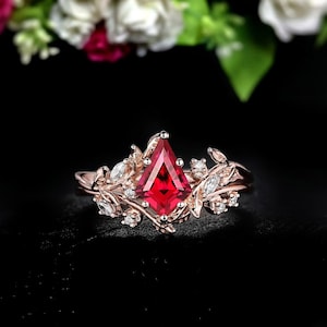 Op de afbeelding: Roségouden ring met een grote, ruitvormige rode edelsteen als middelpunt. De ring is versierd met delicate blad- en rankendetails en kleine, heldere accentstenen. Het ontwerp is geplaatst tegen een zwarte achtergrond.