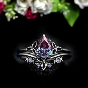 Peut inclure: Un ensemble de bagues en argent foncé avec une pierre précieuse en forme de poire, changeant de couleur. La pierre principale est violet foncé avec des nuances bleues, sertie dans un design filigrané orné. Petites pierres rondes sur la bande.