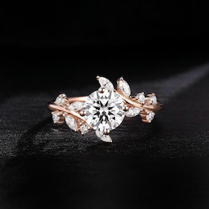 Puede incluir: Anillo de oro rosa con una gema grande, redonda y transparente en el centro, rodeada de gemas más pequeñas y transparentes y detalles en forma de hojas. El anillo se presenta sobre un fondo oscuro, resaltando su intrincado diseño y brillo.