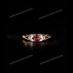 Puede incluir: Un anillo de oro rosa con una piedra central de rubí ovalada, flanqueada por piedras blancas más pequeñas. El anillo tiene un intrincado diseño de filigrana en la banda y alrededor de las piedras. El anillo está sobre un fondo negro.