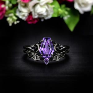 May include: A black ring set featuring a large, coffin-shaped, purple gemstone as the centerpiece. The ring is accented with black crescent moons, black gems, and bat wing designs. The background is black with blurred flowers.