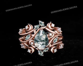 May include: Rose gold ring set featuring a pear-shaped moss agate gemstone. The ring is surrounded by a floral design with small white gemstones and green accents. The set includes a matching band with similar detailing.