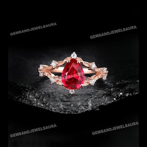 May include: A rose gold ring featuring a pear-shaped, deep red gemstone as the centerpiece. The ring is adorned with small, clear accent stones and a decorative, openwork design. The ring is set against a dark, textured background.