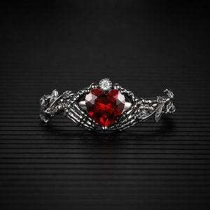 May include: A silver ring featuring a heart-shaped, faceted, red gemstone held by skeletal hands. A small, round, clear gemstone sits above the heart. The ring has a detailed, floral design on the band.