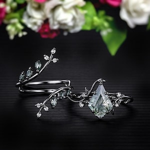 Könnte beinhalten: Zwei Ringe mit einem von der Natur inspirierten Design. Ein Ring hat einen großen, hellgrünen, rautenförmigen Stein. Beide Ringe haben ein dunkles Metallband mit kleinen grünen Steinen und klaren Kristallen.