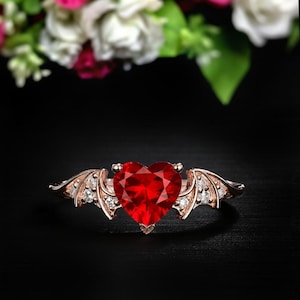 May include: A rose gold ring featuring a faceted, heart-shaped red gemstone. The ring has small white gemstones and wing-like designs on either side of the heart. The ring is set against a black background.