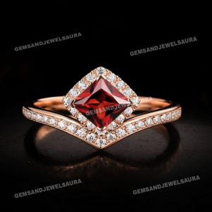 May include: A rose gold ring featuring a square, deep red gemstone at its centre, encircled by a halo of small, clear stones. The band has a V-shaped design, also embellished with small, clear stones. The ring is set against a black backdrop.