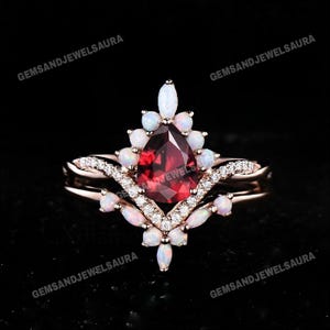 Może przedstawiać: Pierścionek z różowego złota z gruszkowym, ciemnoczerwonym kamieniem szlachetnym pośrodku, otoczonym białymi opalowymi akcentami i małymi, przezroczystymi kamieniami. Pierścionek ma podwójną obrączkę. Widoczny tekst "GemsandJewelsaura".