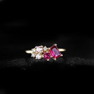 May include: A gold ring featuring a cluster of gemstones. The ring has a large, rectangular, deep pink gemstone, surrounded by smaller pink and white stones. The ring is set against a black background.
