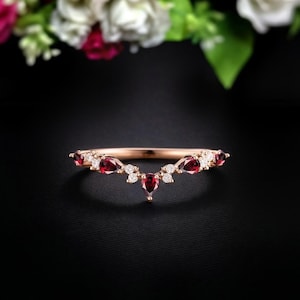 May include: Rose gold band ring with a curved design. The ring features pear-shaped red gemstones and small round white gemstones. The ring is set against a black background with a blurred floral arrangement in the background.