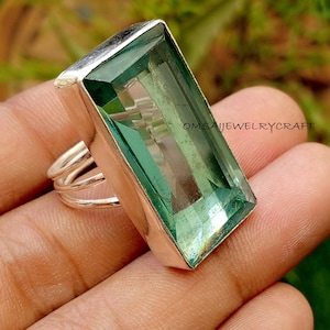 May include: A silver ring featuring a large, rectangular, faceted green gemstone set in a smooth metal bezel. The band consists of three thin, parallel silver wires. The text OMSAIJEWELRYCRAFT is visible on the surface of the green stone.