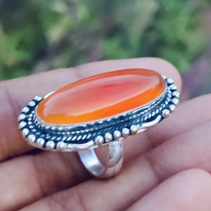 Puede incluir: Un anillo de plata con una piedra preciosa ovalada de color rojo anaranjado. La piedra preciosa está engastada en un engaste de plata detallado y con cuentas. El anillo tiene un acabado pulido y una banda sencilla. El texto "OMSAJEWELRYCRAFT" es visible.