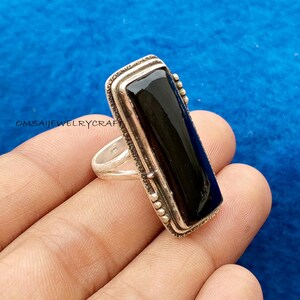 May include: A rectangular silver ring featuring a polished black stone. The ring has a detailed, textured border. The image is set against a blue backdrop. The text "OMSAIJEWELRYCRAFT" is visible.